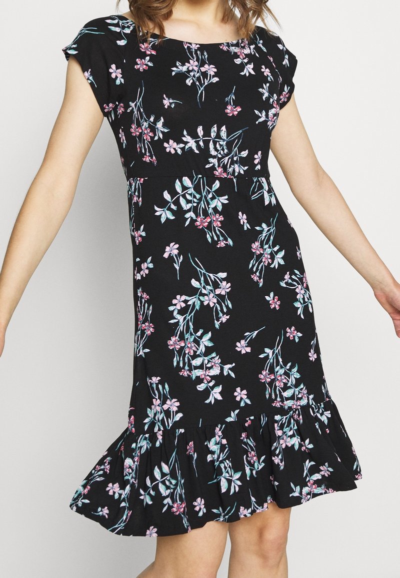 Femme portant une robe noire mi-longue avec un motif floral rose et blanc et un ourlet à volants, debout les bras légèrement étendus.