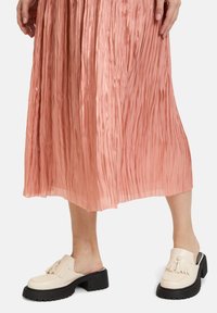 Scarpe beige slip-on con tomaia texture e dettagli frangi, abbinate a una gonna plissettata rosa; le scarpe hanno una suola nera chunky.