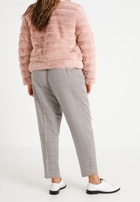 Giacca in pelliccia sintetica rosa chiaro con righe orizzontali, abbinata a pantaloni a quadri in nero, bianco e rosso, e scarpe bianche.