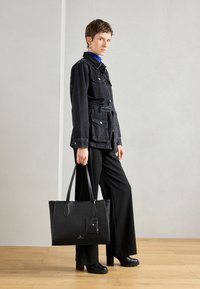 Geantă tote neagră, structurată, din piele netedă, cu mânere duale, un tag și detalii subtile cu logo. Asortată cu o jachetă din denim și pantaloni negri.