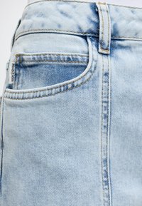 Φωτεινό γαλάζιο τζιν κοντινή λήψη που δείχνει την μπροστινή τσέπη, θηλιά ζώνης και ραμμένη ετικέτα με την επιγραφή «Modström» στην άκρη της τσέπης.