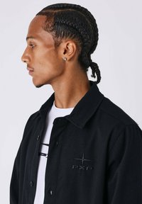 Zwarte button-up jas in een gestructureerde stof, met een geborduurd logo op de linkerborst en een klassiek kraagontwerp.