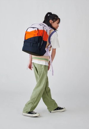 Backpack in purple, orange, and navy with multiple compartments. Textured fabric, adjustable straps, and a sturdy base. Model wears loose green pants.