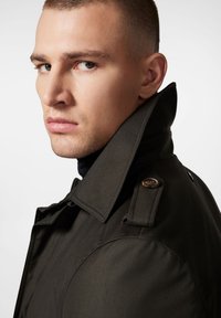 Ciemnozielony płaszcz trencz z teksturowanej tkaniny, z wysokim kołnierzem i epoletami. Posiada metalowy detal z guzikami w okolicy ramienia.