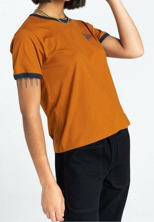 Persona que lleva una camiseta de manga corta naranja con ribete oscuro y pantalones negros, un brazo levantado y un tatuaje visible en la parte superior del brazo.