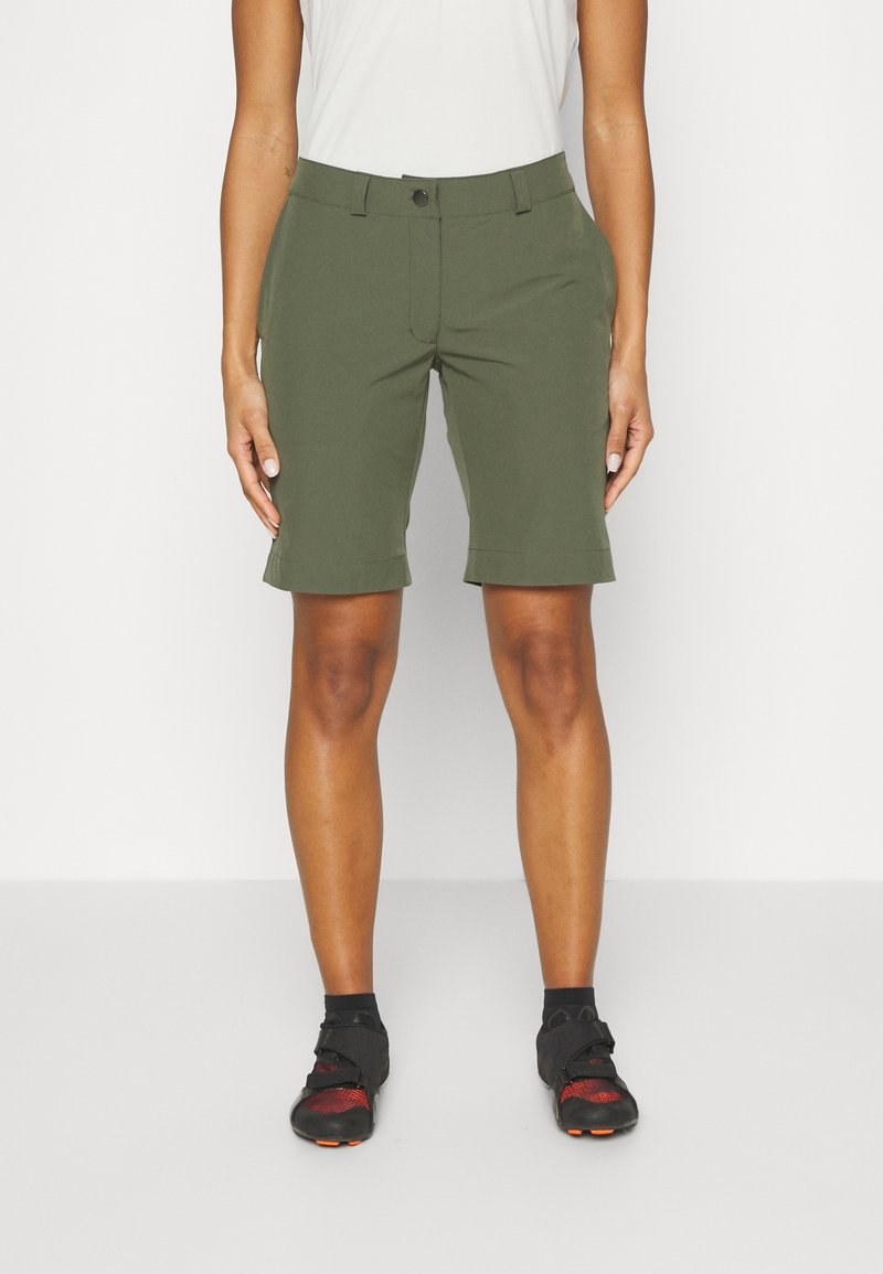Personne portant un short vert olive jusqu'aux genoux, un haut blanc, des chaussettes noires et des chaussures de cyclisme noires et rouges, debout devant un fond uni.
