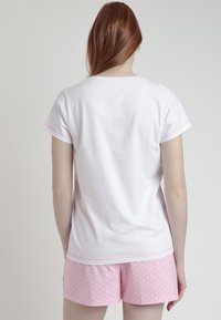 Mujer con el cabello largo y rojo, vistiendo una blusa blanca de manga corta y unos pantalones cortos rosas con lunares, de pie de espaldas contra un fondo liso.