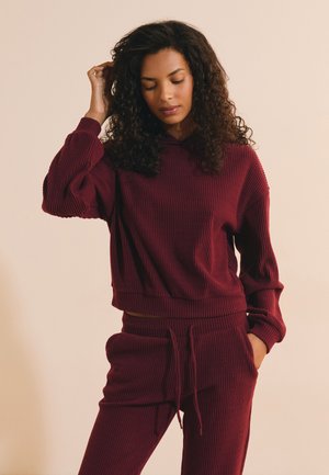 Femme aux cheveux bouclés portant un sweat à capuche côtelé bordeaux et un pantalon assorti, debout, une main touchant ses cheveux et les yeux fermés.