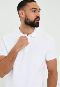 Hvid polo-shirt lavet af blødt stof, med en klassisk krave og korte ærmer. Et lyseblåt logo er broderet på venstre bryst.