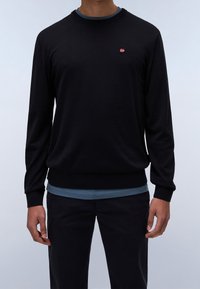 Uomo che indossa un maglione nero a maniche lunghe, senza fantasia, con un piccolo logo di una bandiera rossa, bianca e blu sul petto, sopra una camicia blu.