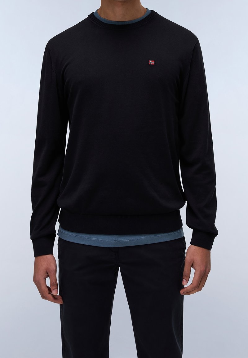 Uomo che indossa un maglione nero a maniche lunghe, senza fantasia, con un piccolo logo di una bandiera rossa, bianca e blu sul petto, sopra una camicia blu.