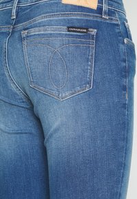Vaqueros de mezclilla azul oscuro con un diseño de cinco bolsillos, que presentan una etiqueta visible de Calvin Klein y costuras en contraste en los bolsillos traseros.