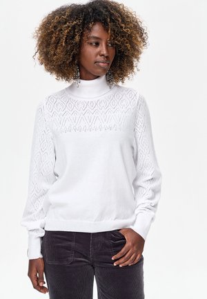 Witte gebreide trui met een hoge hals, voorzien van een ruitpatroon op het beleg en gestructureerde mouwen. Gecombineerd met donkergroene ribfluwelen broek.