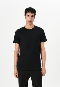 Tricou negru cu mânecă scurtă, confecționat dintr-un material moale. Are un guler rotund și o croială relaxată, purtat cu pantaloni negri, pe un fundal simplu.
