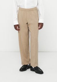 Beige maßgeschneiderte Hose mit glatter Textur und elastischem Bund, kombiniert mit schwarzen Slippern und getragen mit einem weißen Hemd mit Knöpfen.