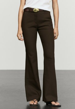 Kobieta w ciemnobrązowych rozszerzanych spodniach z czarnym paskiem i białym topem stoi na gładkiej podłodze w czarnych sandałach.