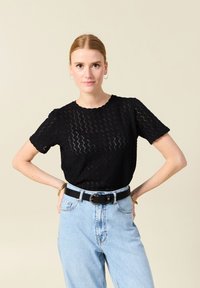Schwarzes Kurzarmoberteil mit strukturiertem Zickzackmuster, rundem Halsausschnitt und gewelltem Rand, kombiniert mit hochgeschnittenen hellblauen Jeans und einem schwarzen Gürtel.