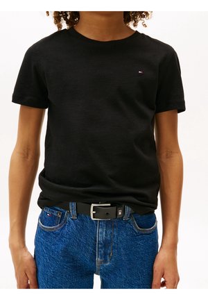 Person trägt ein schwarzes T-Shirt mit kurzen Ärmeln und blaue Jeans mit schwarzem Gürtel, kleines Logo auf dem Shirt und Gürtel-Schnalle sichtbar.