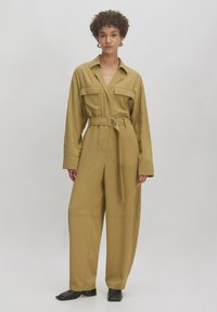 Helles olivgrünes Jumpsuit mit tailliertem Gürtel, großen Brusttaschen und weitem Bein. Hergestellt aus glattem, leicht glänzendem Stoff.
