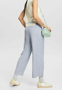 Kobieta z warkoczami, ubrana w bezrękawnik z dzianiny, jasnoniebieskie szerokie spodnie, białe sneakersy, szare skarpetki i nosząca miętową torbę na ramię.