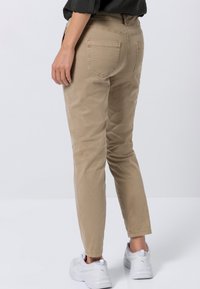 Chinos beiges avec cinq poches, jambes fuselées et texture subtile. Portés avec des baskets blanches. Vue arrière montrant les détails de couture.