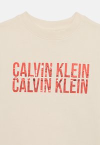 Beige mikina so zvýrazneným červeným nápisom "CALVIN KLEIN" opakovaným po celej ploche, s hladkou textúrou a okrúhlym výstrihom.