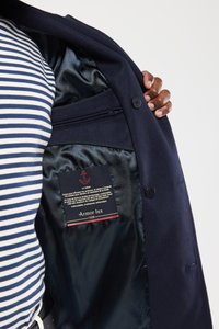 Granatowy wełniany płaszcz z jedwabną podszewką, z metką z logo kotwicy, a częściowo widoczna jest paskowana koszula.