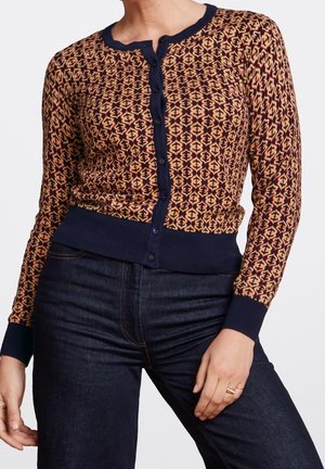 Vrouw draagt een cardigan met lange mouwen en knopen, voorzien van geometrische patronen in rood, geel en marineblauw, gecombineerd met donkerblauwe spijkerbroek en een gouden ring aan de linkerhand.