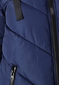 Granatowa kurtka puchowa z ukośnym wzorem pikowania, zamkiem błyskawicznym z przodu i czarnymi akcentami na elementach metalowych. Miękka w dotyku i ocieplona dla komfortu cieplnego.