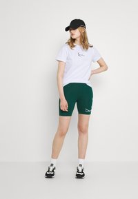 Hvit t-skjorte med svart logo, kombinert med grønne sykkelshorts med hvit linjedesign. En svart caps og sorte joggesko fullfører antrekket.