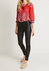 Röd genomskinlig blus med paisleymönster, elastiska manschetter; kombinerad med svarta åtsittande leggings. Modellen bär espadriller med virkad detalj.