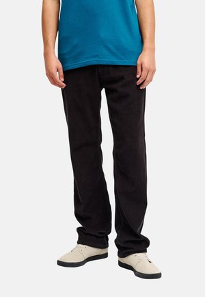 Person, die schwarze Cordhosen, beige Schuhe und ein blaues Hemd trägt, steht vor einem einfarbigen hellen Hintergrund.
