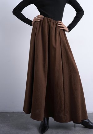 Kvinne iført en høytlivs, teksturert brun maxiskjørt med svarte spisse ankelstøvler og en tettsittende svart langermet topp, hendene på hoftene.