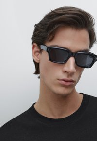 Schwarze quadratische Sonnenbrille mit dunklen Gläsern und grauen Akzenten, getragen von einer Person in einem schwarzen T-Shirt. Glatte Oberfläche mit minimalen Details.