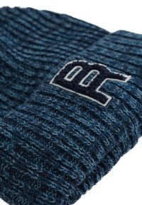 Blå stickad mössa med ett texturerat mönster, med en mörkblå mudd som har en vit broderad logotyp i mitten.