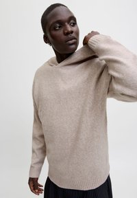 Suéter de punto texturizado en marrón claro, con capucha y puños acanalados, que se lleva sobre pantalones a rayas negros. Material suave con un ajuste relajado.