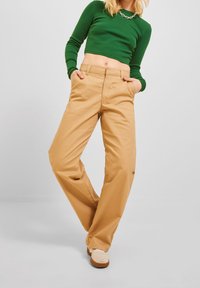 Beige weiter Hosen mit glatter Textur, ausgestattet mit Seitentaschen und flachem Vorderteil. Kombiniert mit einem grünen Langarmshirt.