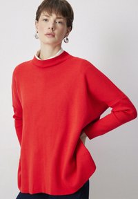 Crvena, oversized veste s rebrastim visokim vratom i rukavima. Mekana tekstura, opušteni kroj i drapirani dizajn. Nosi se preko bijele košulje s kragnom.