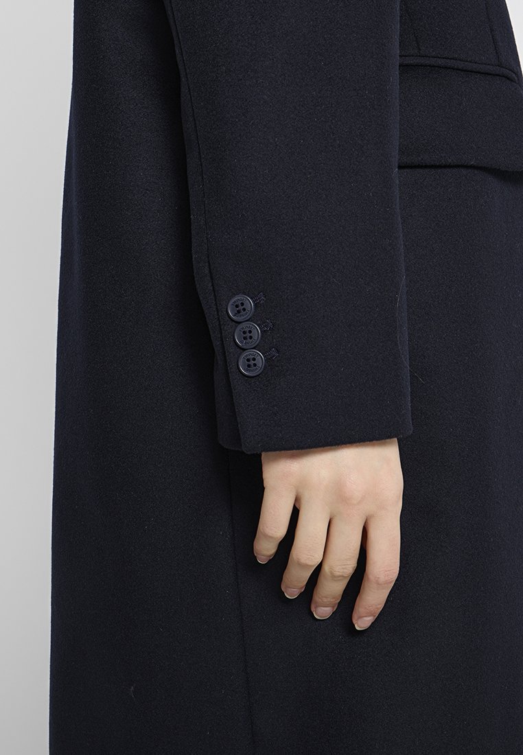 Navy wollen jas met een mouw met vier donkerblauwe knopen. De stof lijkt glad met een licht gestructureerde afwerking. Tailored fit, minimalistisch ontwerp.