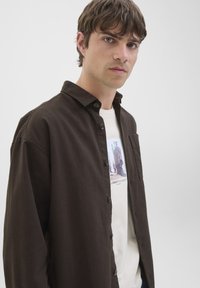 Joven con cabello corto y castaño que lleva una camisa de botones marrón oscuro sobre una camiseta blanca estampada, mirando hacia adelante contra un fondo liso.