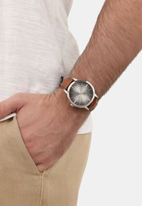 Braunes Lederarmbanduhr mit silbernem Gehäuse, grauem Zifferblatt, goldenen Stundenmarkierungen und minimalistischer Gestaltung; Hand ruht in einer khakifarbenen Tasche.