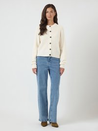 Mulher em pé, vestindo um suéter creme com botões, calças jeans de perna larga azul claro e botas de salto castanhas, contra um fundo claro e simples.