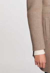 Abrigo de lana color marrón claro con un acabado texturizado, bolsillo lateral y mangas de punto en contraste color crema. Presenta una forma suave y ajustada.