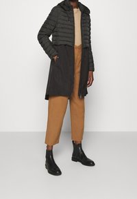 Abrigo negro acolchado con sección superior ajustada y parte inferior más larga. Llevado sobre una camiseta beige y pantalones marrones cortos, combinado con botines negros.