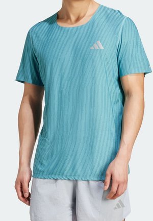 Mann trägt hellblaues strukturiertes Sport-T-Shirt und hellgraue Shorts, steht mit entspannten Armen vor einfachem Hintergrund.