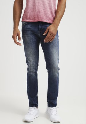 Dark blue denim jeans with a slim fit, featuring subtle fading on the thighs, paired with a short-sleeved pink T-shirt and white sneakers.