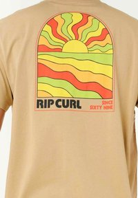 Tan katoenen t-shirt met een kleurrafische afbeelding van golvende lijnen in oranje, geel en groen, en de tekst "RIP CURL SINCE SIXTY NINE" onderaan.
