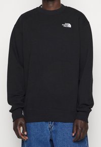 Musta collegepaita, valmistettu puuvillasekoitteesta, jossa on pyöreä pääntie, pitkät hihat, resorit hihansuuissa ja pieni valkoinen logo vasemmassa rinnassa.