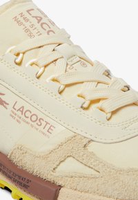 Zapatilla Lacoste en amarillo pálido con detalles en ante. Presenta paneles texturizados, cordones planos y branding en el lateral y la lengüeta. Suela de goma.