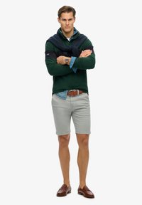 Groen overhemd met lange mouwen, gedragen over een lichtblauw denim shirt, gecombineerd met grijze shorts en brown loafers; beschikt over een marineblauwe trui om de hals geknoopt.